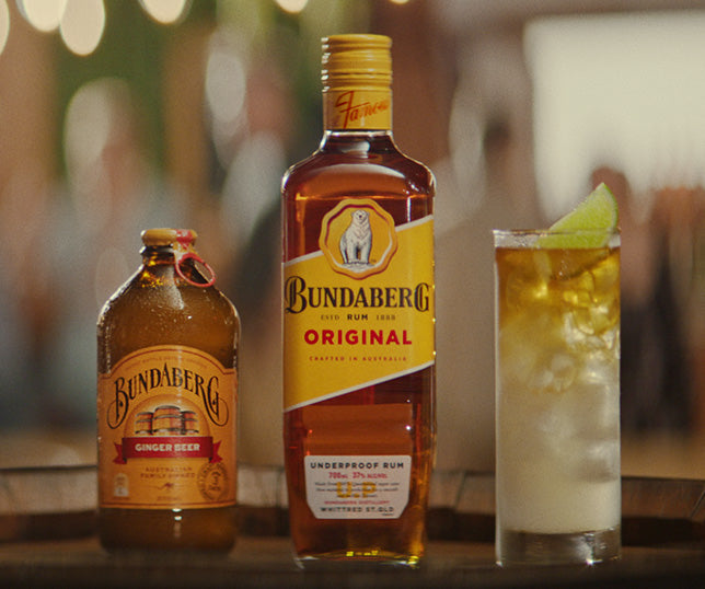 A Bundaberg ginger beer bottle, a Bundaberg Original rum bottle, and an ice-filled highball with a lime wedge sit on a wooden bar counter with warm, blurred lights behind. Transcribed text: - Bundaberg - Ginger Beer - Bundaberg - ORIGINAL - UNDERPROOF RUM