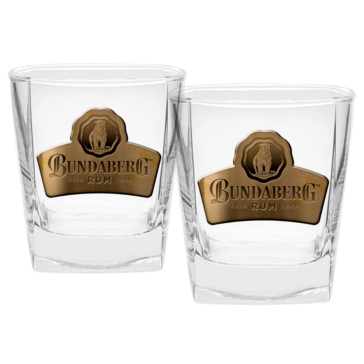Two clear short whiskey glasses sit side-by-side on a white background, each bearing a gold oval label with a crest; the small printed logo/text on the labels is too small to read.
