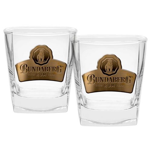 Two clear short whiskey glasses sit side-by-side on a white background, each bearing a gold oval label with a crest; the small printed logo/text on the labels is too small to read.