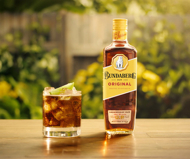 A bottle of Bundaberg rum stands beside a short glass of rum and ice garnished with a lime wedge, on a wooden table in a sunlit backyard garden.

Text on bottle: "Bundaberg" "Original" "Underproof Rum"
Small fine print (alcohol % and other small labels) is too small to read.
