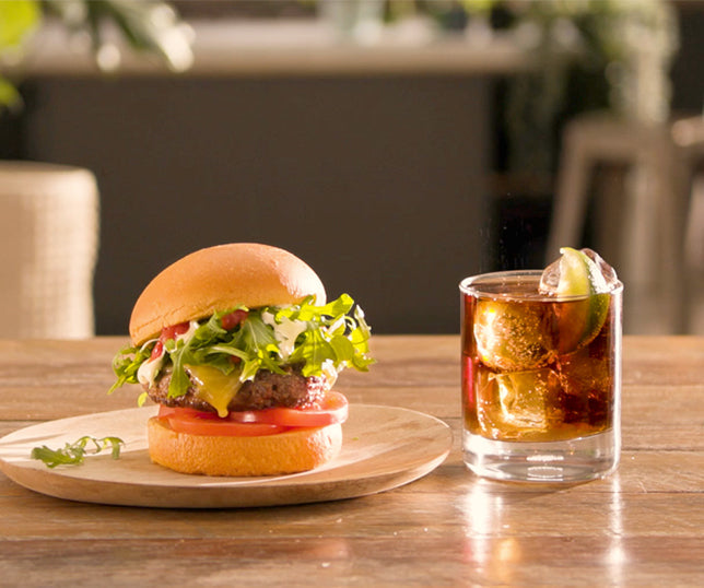 A cheeseburger rests on a wooden plate with lettuce, tomato, melted cheese and sauce; a glass of cola with ice and lime sits beside it on a table in sunlight.