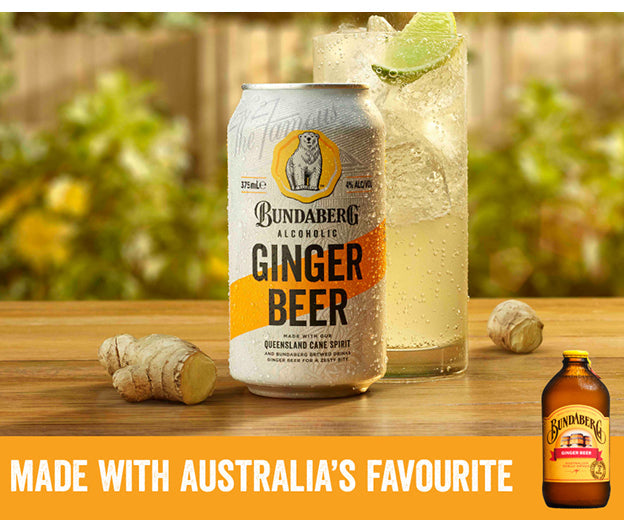 A Bundaberg canned ginger beer sits centered on a wooden table, flanked by a tall ice-and-lime glass and pieces of fresh ginger, set against a blurred backyard garden.