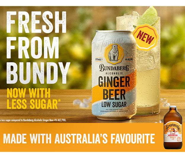 Chilled can of Bundaberg Low Sugar Ginger Beer sits on a wooden table beside an iced lime drink, against a blurred leafy background with a small Bundaberg bottle at bottom-right.