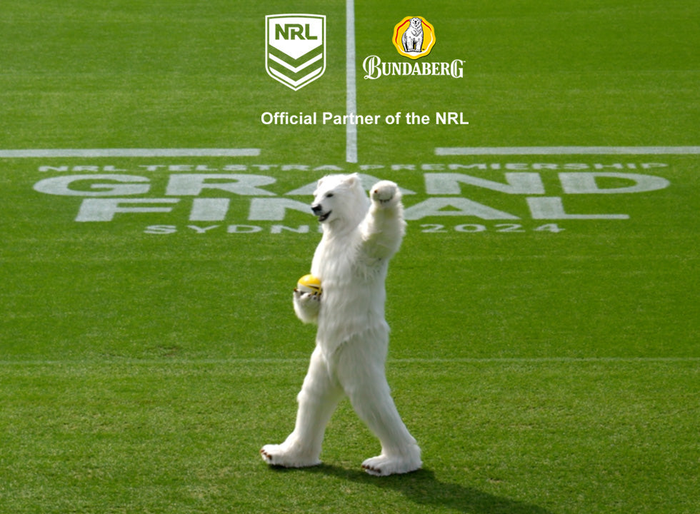Polar bear mascot walking and waving while holding a yellow ball on a grassy stadium field with painted text "GRAND FINAL SYDNEY 2024"; visible logos/text: "NRL", "Bundaberg", "Official Partner of the NRL".