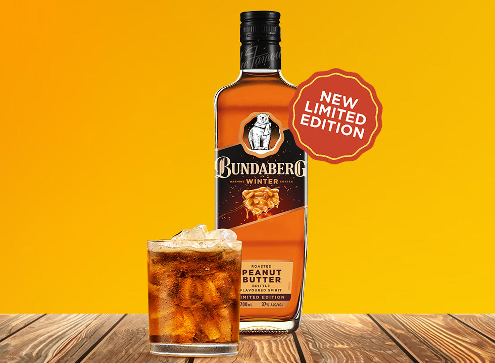 Bundaberg bottle labeled as a limited-edition peanut-butter brittle spirit stands behind an iced tumbler on a wooden table, set against a solid yellow background.