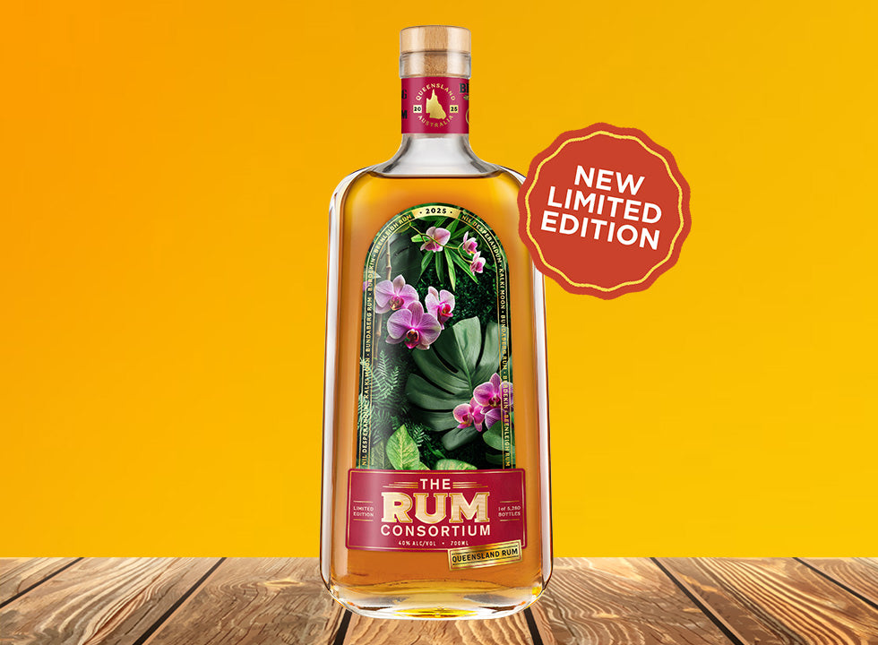 A tall glass bottle of rum stands upright on a wooden tabletop, showing a tropical floral label; bright yellow background and a red scalloped badge hover to the bottle's right.