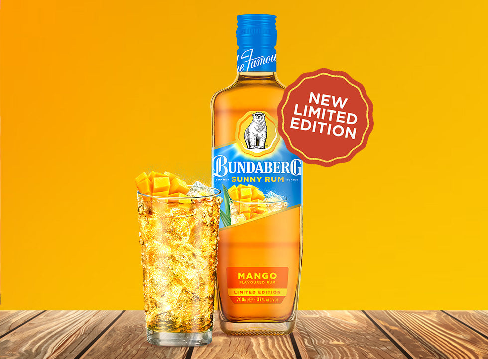 A Bundaberg rum bottle stands beside an ice-filled mango cocktail on a wooden surface against a bright yellow background.