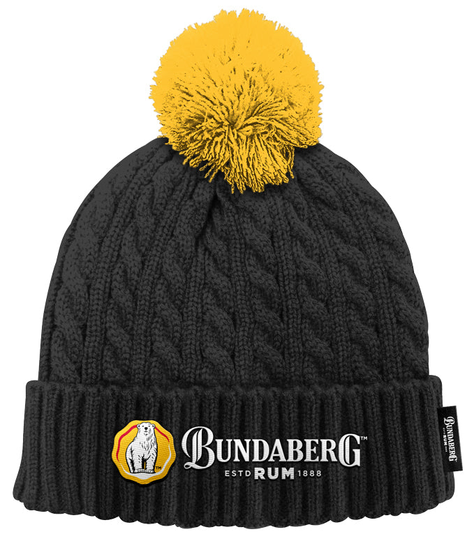Black cable-knit beanie with a yellow pom-pom rests on a white background; folded cuff displays an embroidered Bundaberg Rum logo with circular seal.