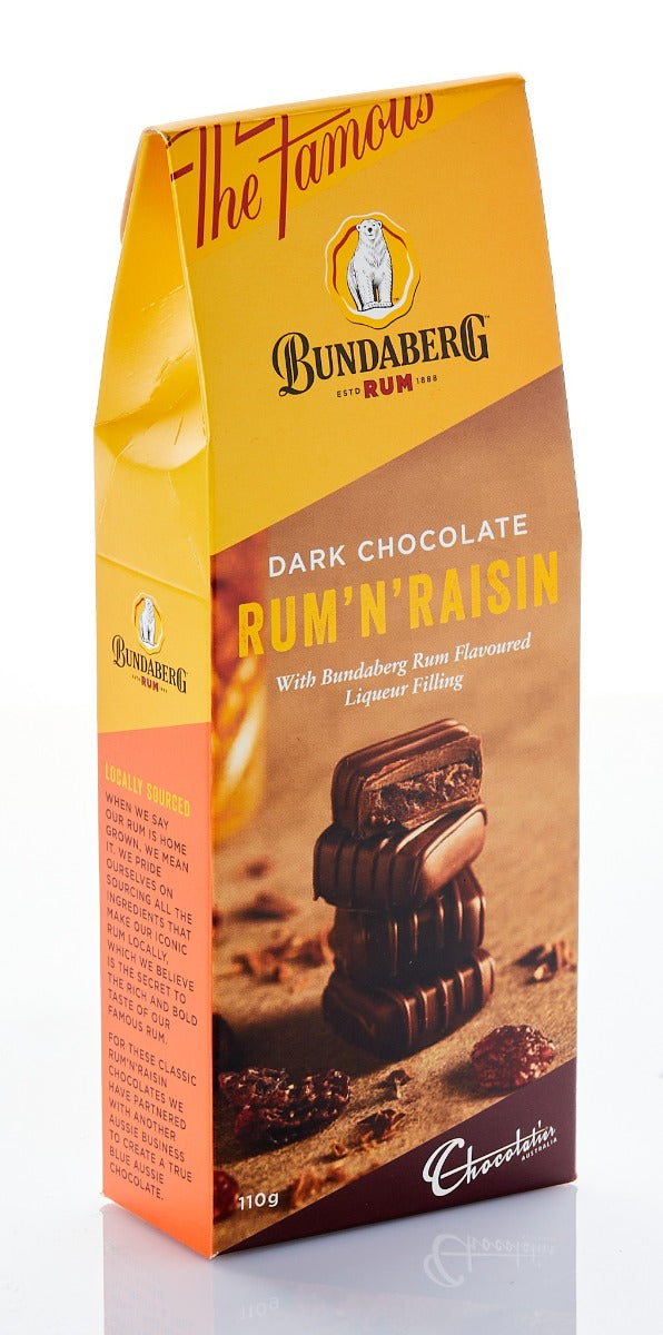 Tall yellow-and-brown chocolate box stands upright on a reflective white surface, showing stacked dark chocolate squares and raisins; studio-lit, viewed slightly from the front-right.
