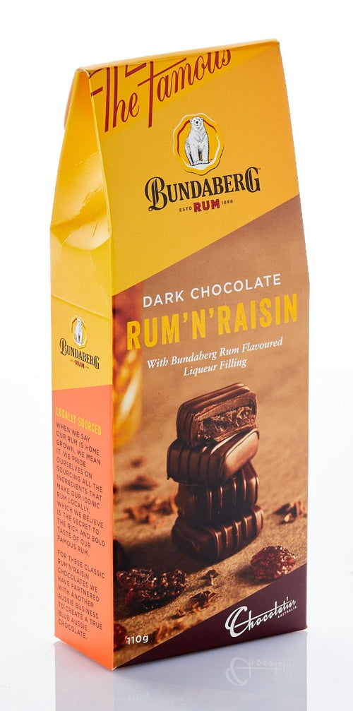 Tall yellow-and-brown chocolate box stands upright on a reflective white surface, showing stacked dark chocolate squares and raisins; studio-lit, viewed slightly from the front-right.