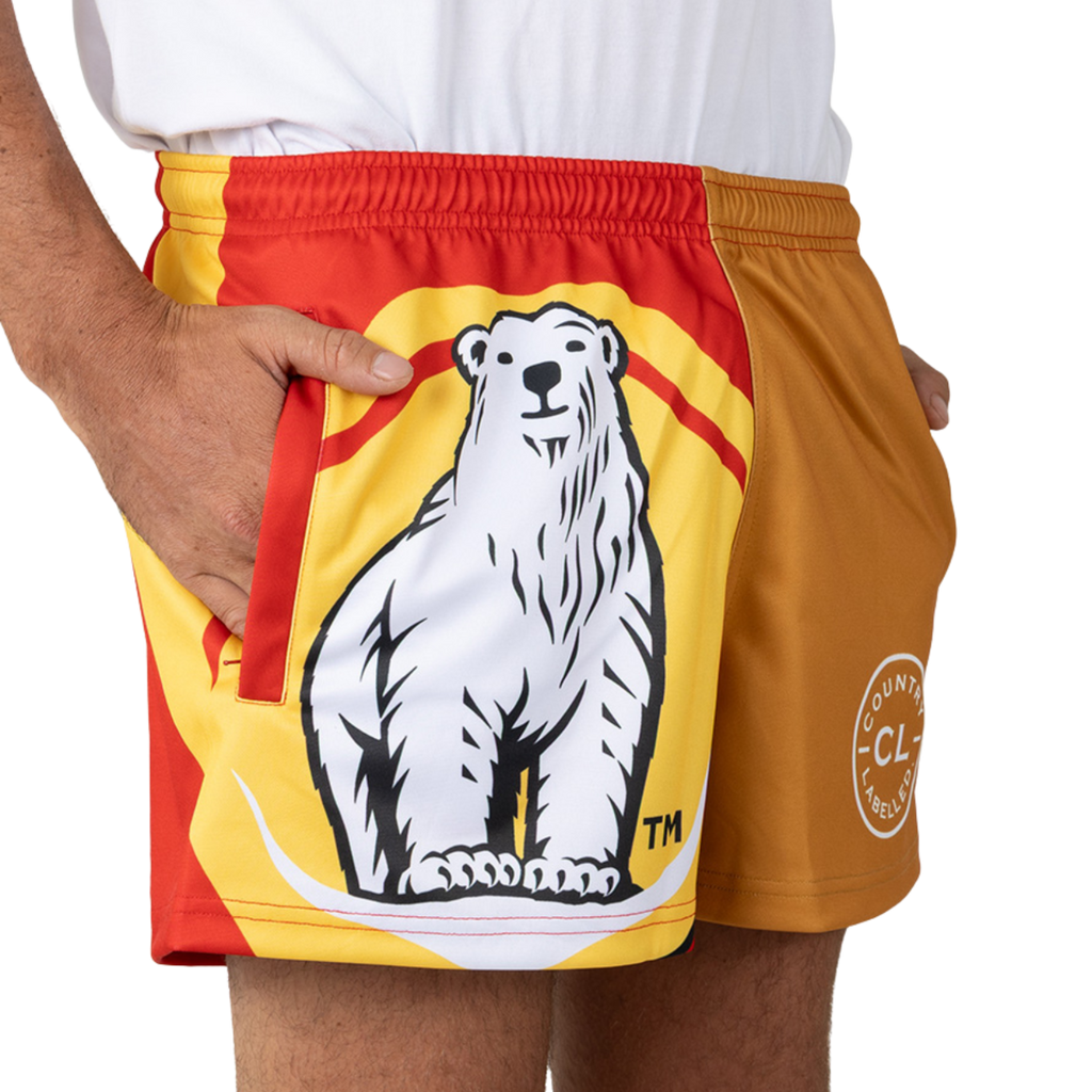 Shorts featuring a large illustrated polar bear on the front; person stands cropped at the hips with one hand in a pocket, wearing a white shirt and red-yellow-orange sport shorts on a white background.