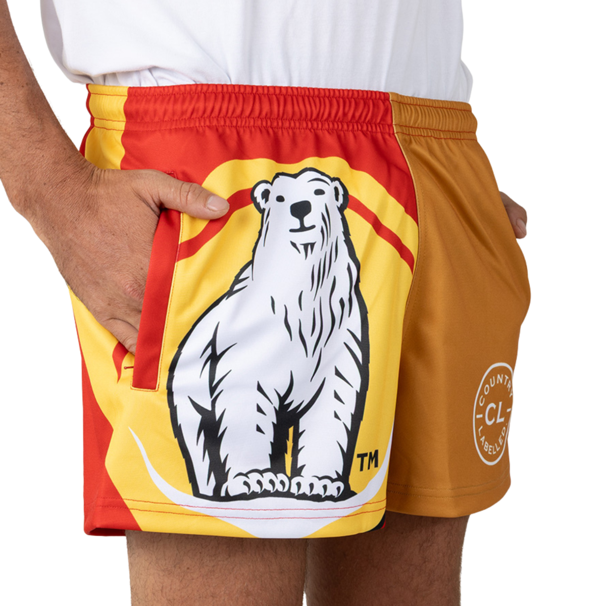 Shorts featuring a large illustrated polar bear on the front; person stands cropped at the hips with one hand in a pocket, wearing a white shirt and red-yellow-orange sport shorts on a white background.