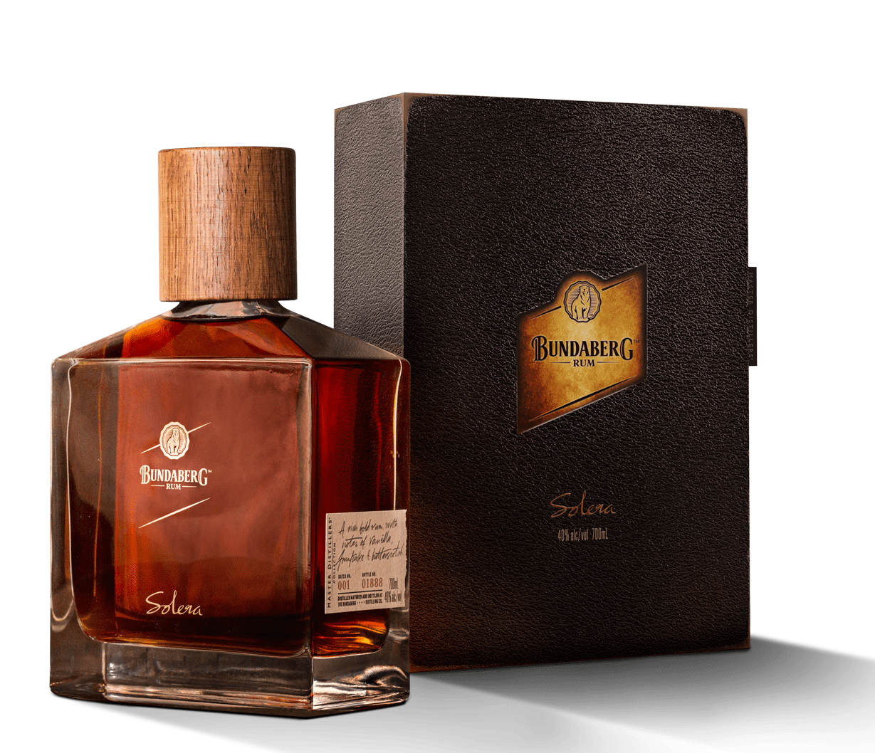 Amber whisky bottle with wooden stopper sits beside a textured black gift box; both rest on a white surface with soft studio lighting and cast distinct shadows. Text on labels is too small/blurred to read—please provide a higher-resolution or close-up image for full transcription.