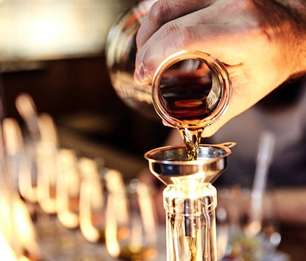A hand tilts a glass decanter, pouring amber liquid through a metal funnel into a clear bottle, set amid blurred rows of similar bottles on a wooden bar.
