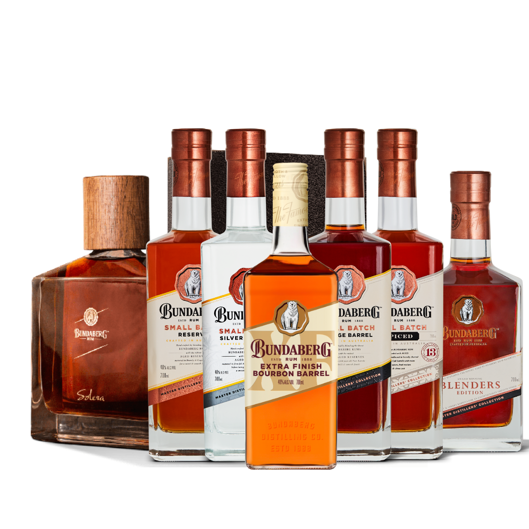 Six assorted spirit bottles arranged in a slight arc, one amber-labeled bottle centered front, a wooden-capped decanter at left, all standing on a white background. Text on labels is too small/blurry to transcribe—please provide a higher-resolution or close-up crop to capture the wording.