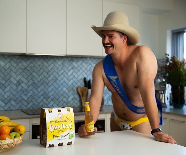 Dingo Dan Leyden holding a bottle of Bundaberg Rum Lazy Bear while wearing a cowboy hat and a pair of Bundaberg Rum Budgy Smugglers inside a kitchen.